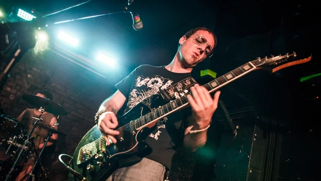 Mężczyzna grający na gitarze elektrycznej podczas koncertu na scenie, intensywne oświetlenie i dynamiczna atmosfera występu.