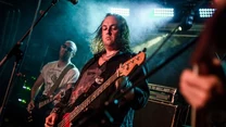 Dwóch muzyków grających na gitarach podczas występu na scenie, skupiona twarz basisty na pierwszym planie, barwne światła sceniczne i sprzęt muzyczny w tle tworzą atmosferę koncertu rockowego.