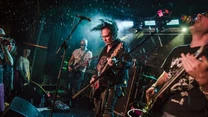 Grupa muzyków energicznie gra na gitarach elektrycznych podczas koncertu na scenie, kolorowe światła sceniczne podkreślają dynamiczną atmosferę występu, a publiczność obserwuje występ z bliska.