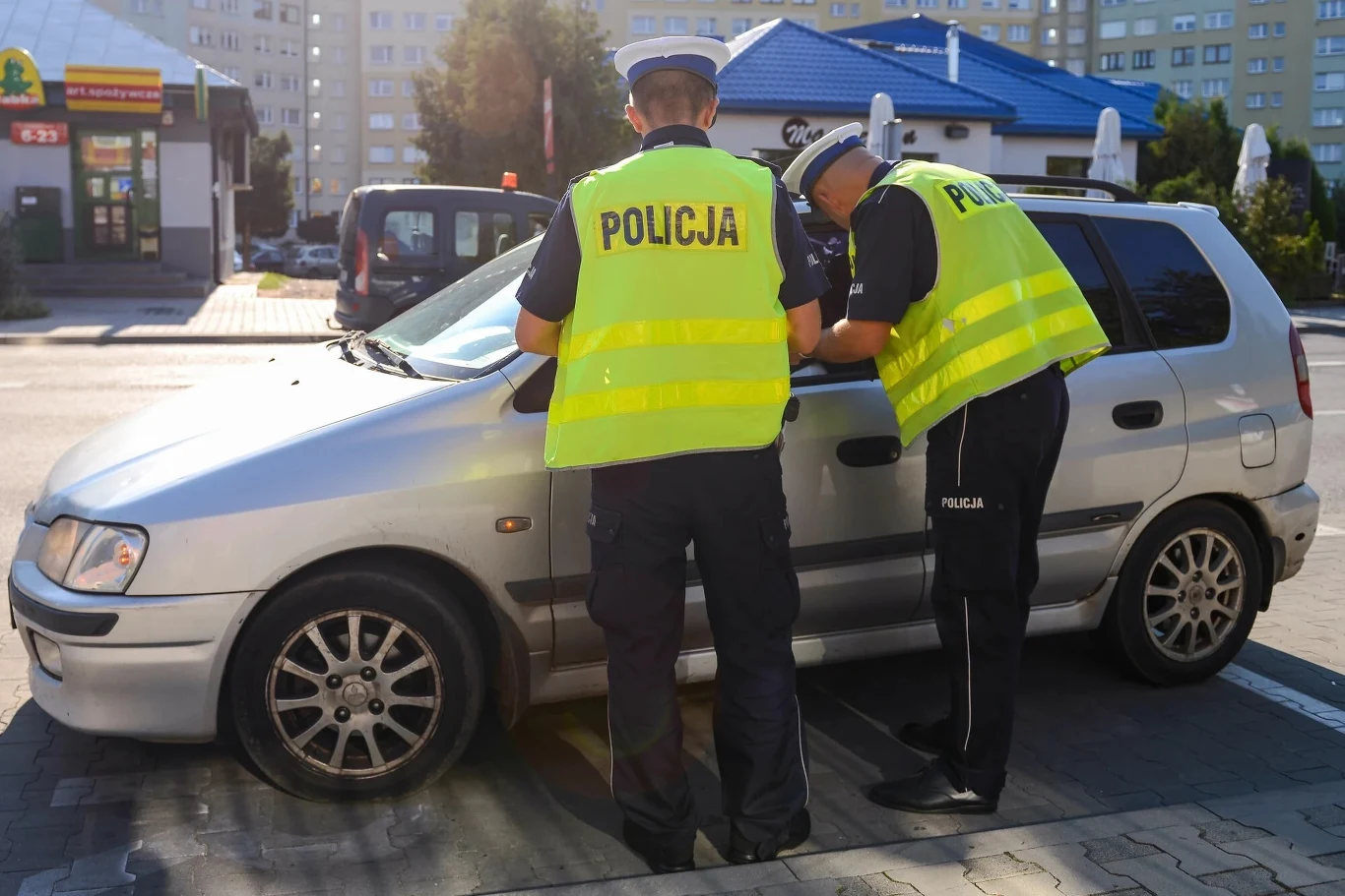 Za korzystanie z nawigacji w telefonie można dostać 500 zł mandatu Dwóch policjantów w żółtych kamizelkach odblaskowych kontroluje srebrny samochód osobowy na miejskim parkingu, spisując notatki przy otwartych drzwiach kierowcy, w tle zabudowa miejska i restauracja.