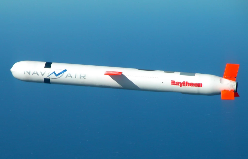 Tomahawk Block IV podczas testów Biała rakieta manewrująca z widocznymi napisami „NAVY AIR” oraz „Raytheon”, wyróżniającymi się czerwonymi elementami statecznika, przemieszczająca się na tle nieba.