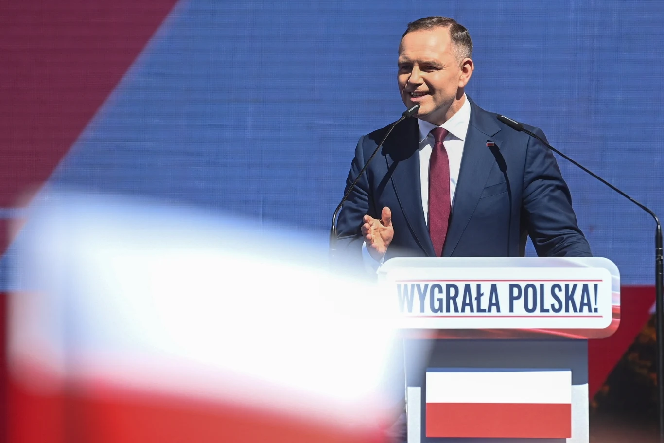 Prezydent elekt Karol Nawrocki przemawiał na terenie amfiteatru w Pułtusku Mężczyzna w garniturze przemawia na scenie podczas wiecu, przed mównicą z napisem 'Wygrała Polska!', w tle duża polska flaga i biało-czerwona flaga na pierwszym planie.