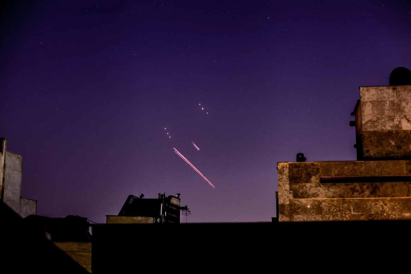 Izraelski atak lotniczy na Teheran oraz obiekty wojskowe w innych miastach Smugi świetlne przecinające nocne niebo nad sylwetkami budynków, światła przypominają tor lotu pocisków lub rakiet, a otoczenie jest spokojne, choć napięte