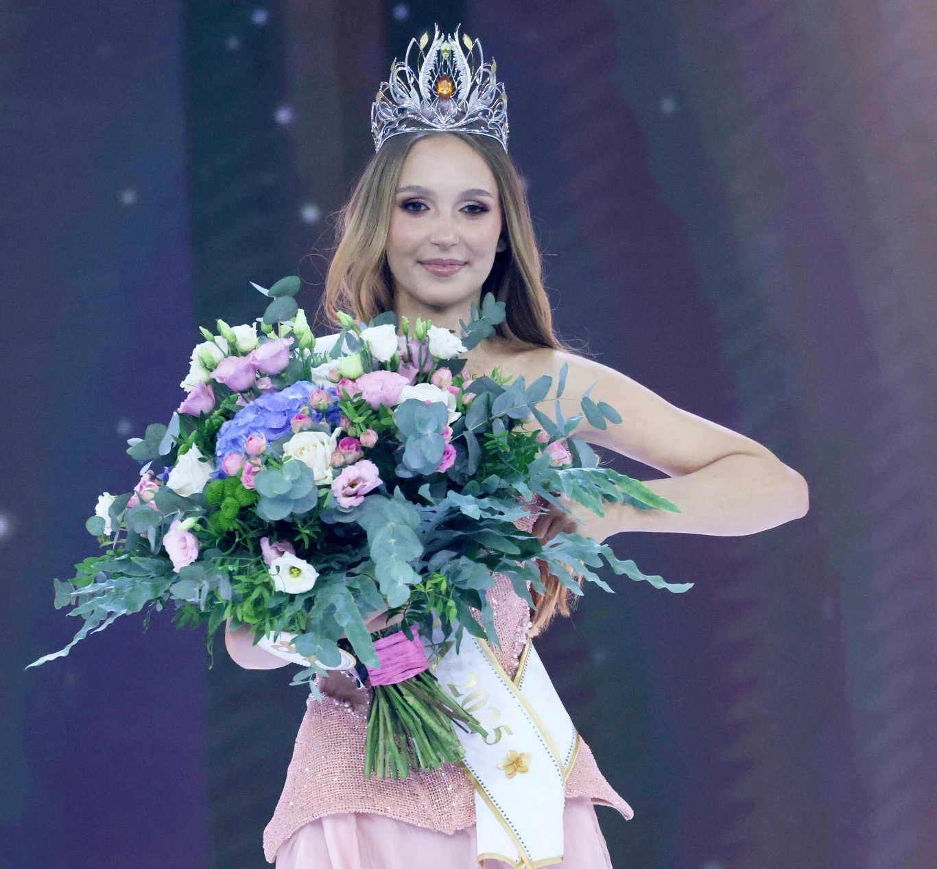 Młoda kobieta z koroną na głowie i szarfą celebruje zwycięstwo na konkursie piękności, trzymając okazały bukiet świeżych kwiatów.