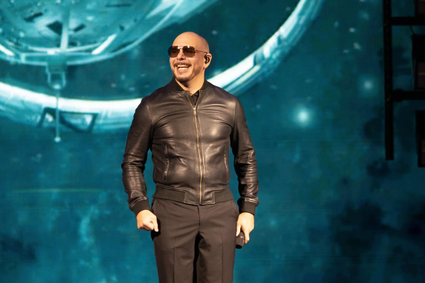 Pitbull zwrócił się po polsku do swoich fanów Mężczyzna w skórzanej kurtce i okularach przeciwsłonecznych stoi na scenie na tle abstrakcyjnej, niebieskiej grafiki przypominającej kosmos lub galaktykę, uśmiechnięty, trzyma w dłoni niewielki przedmiot.