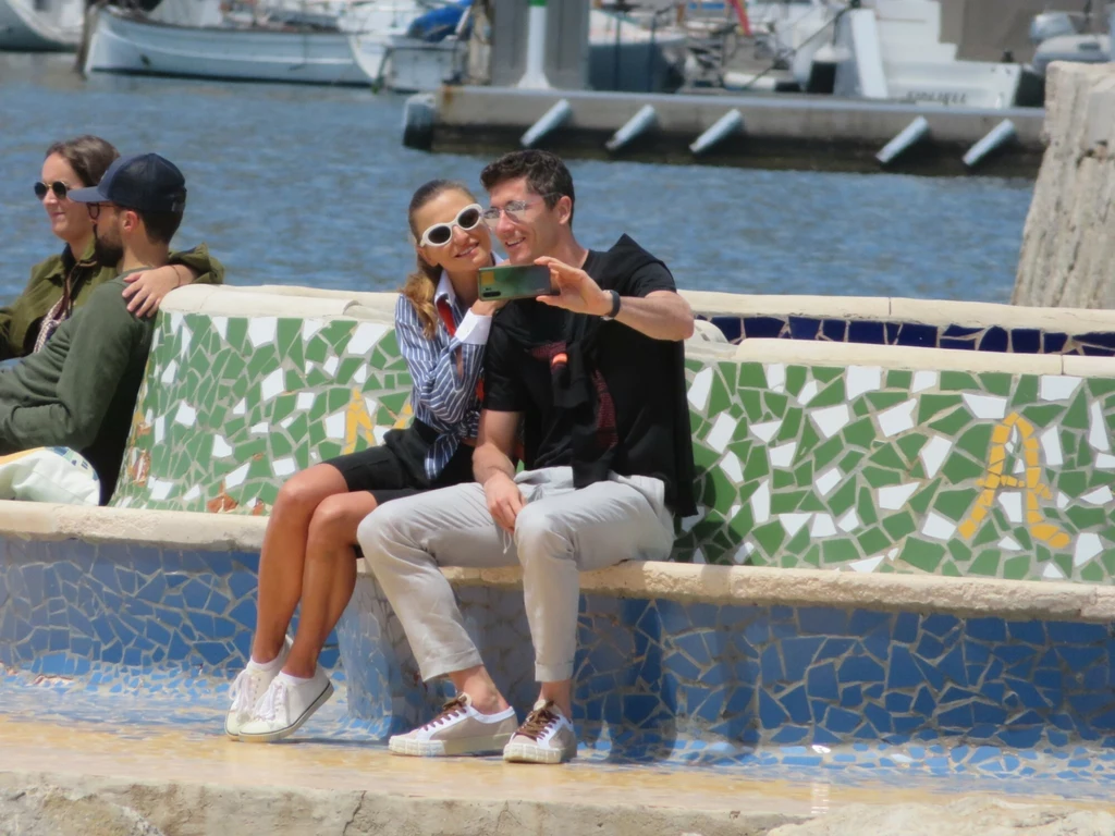 Anna i Robert Lewandowscy w tym roku świętują 12. rocznicę ślubu Para robiąca sobie selfie na kolorowej mozaikowej ławce przy marinie, w tle widać wodę i zacumowane łodzie, obok nich siedzi inna para, wszyscy sprawiają wrażenie zrelaksowanych i pogodnych.