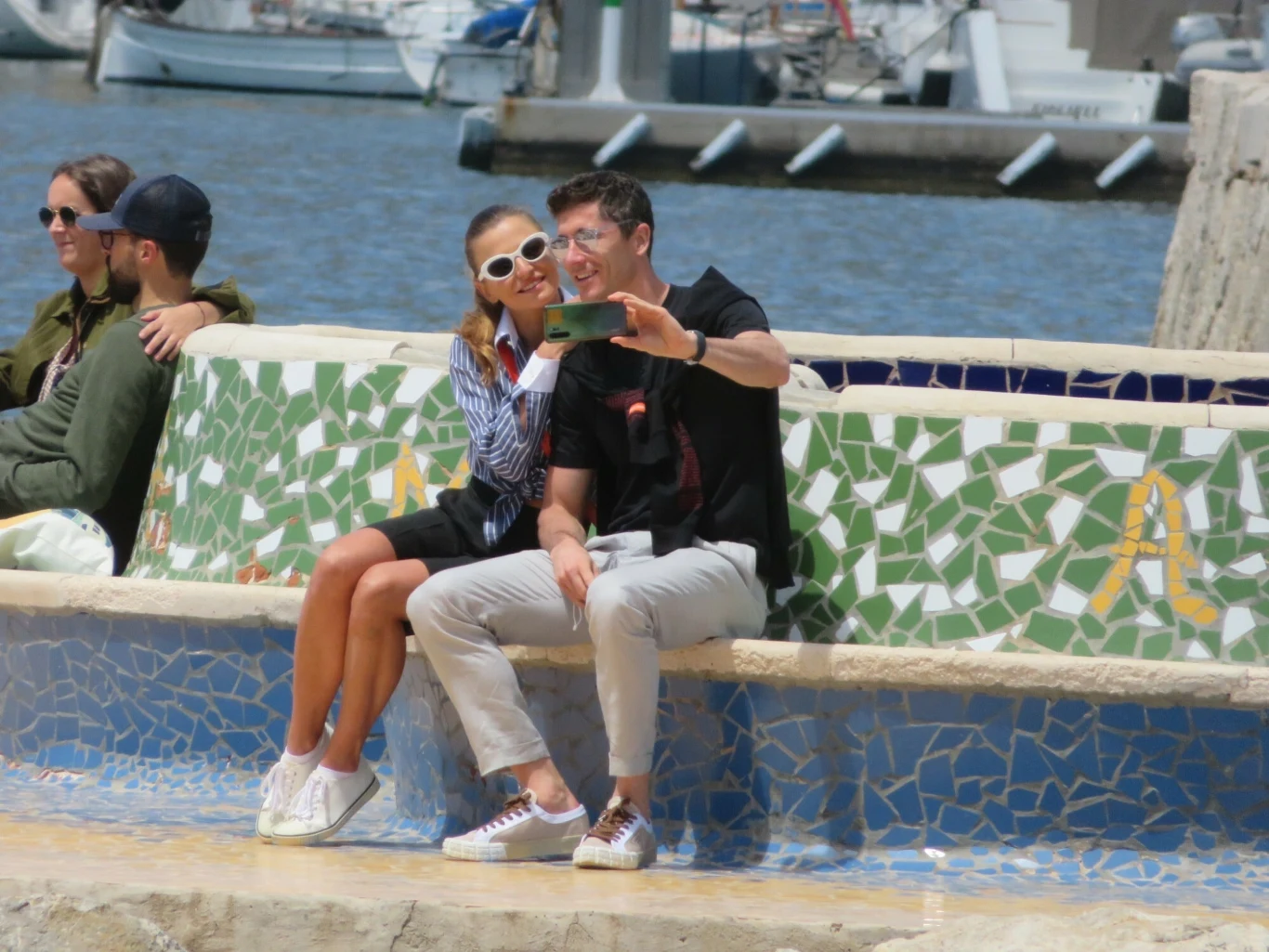 Para robiąca sobie selfie na kolorowej mozaikowej ławce przy marinie, w tle widać wodę i zacumowane łodzie, obok nich siedzi inna para, wszyscy sprawiają wrażenie zrelaksowanych i pogodnych.