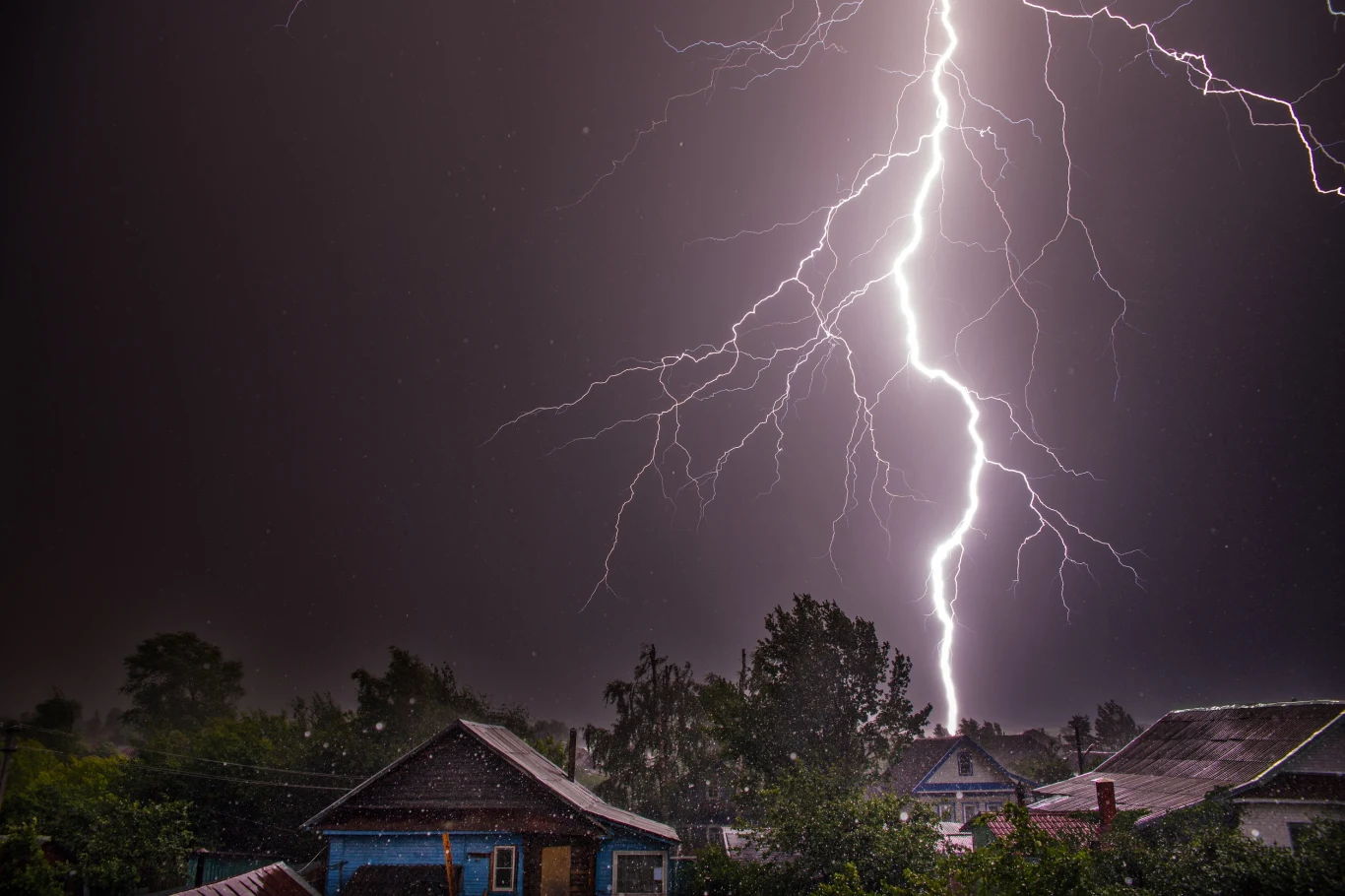 Alert RCB z powodu niebezpiecznych burz obejmuje 16 województw. Niemal cała Polska będzie sią zmagać z groźnymi zjawiskami. Zdjęcie ilustracyjne Jasny piorun uderza z nieba w pobliżu domów, rozświetlając nocne niebo i rzucając ostre światło na pobliską zabudowę i drzewa.