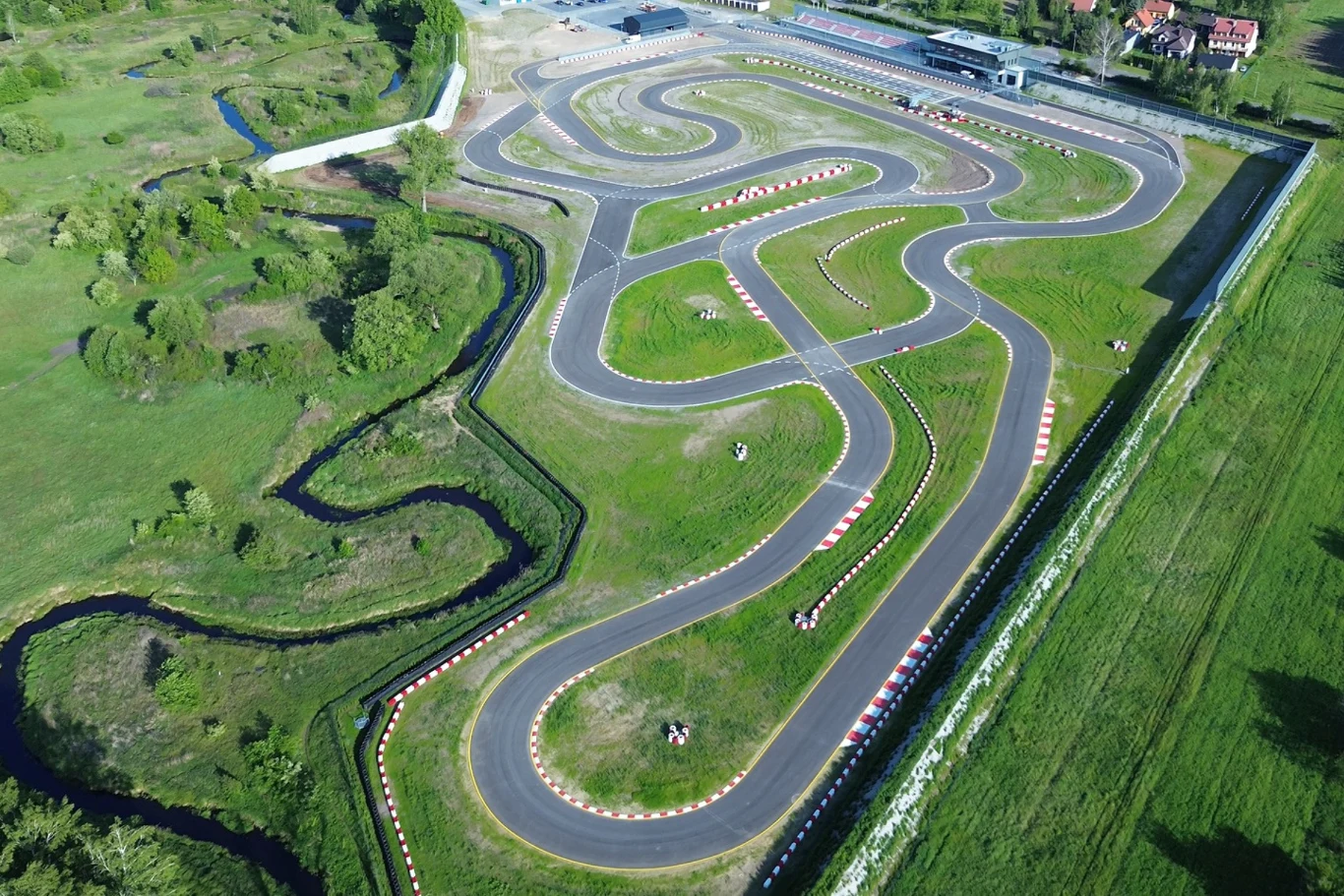 Kręty tor wyścigowy otoczony zielonymi terenami, z zaznaczonymi pobocznymi barierkami i infrastrukturą w tle, idealny do jazdy gokartami lub treningów sportów motorowych.