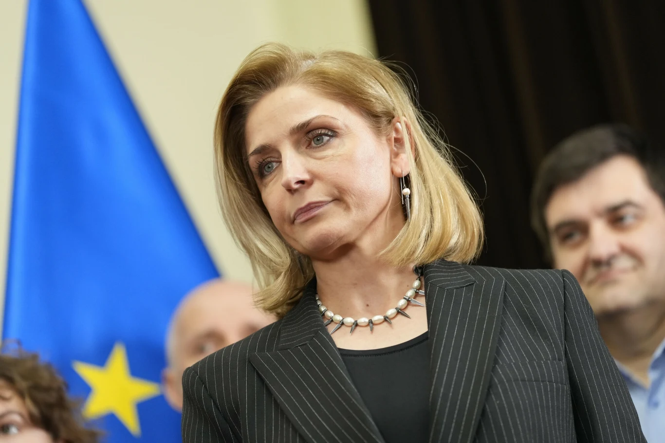 Joanna Mucha podała się do dymisji Kobieta w garniturze z perłowym naszyjnikiem stoi na tle flagi z żółtą gwiazdą i niebieskim tłem, w otoczeniu kilku osób, patrząca poważnym wzrokiem w stronę kamery.