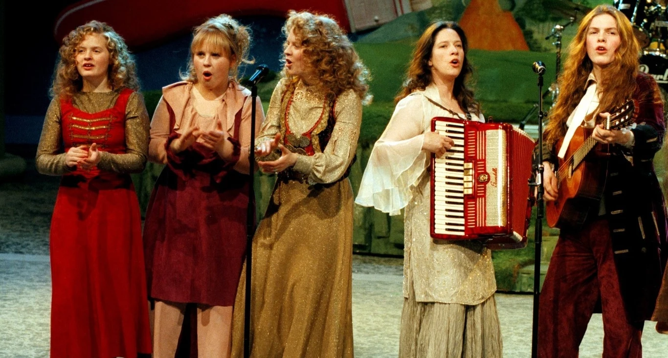 Barby (z lewej) z The Kelly Family Pięć kobiet w strojach scenicznych śpiewa wspólnie na scenie, jedna z nich gra na akordeonie, a druga na gitarze, wszystkie stoją blisko siebie, wyrażając emocje podczas występu.
