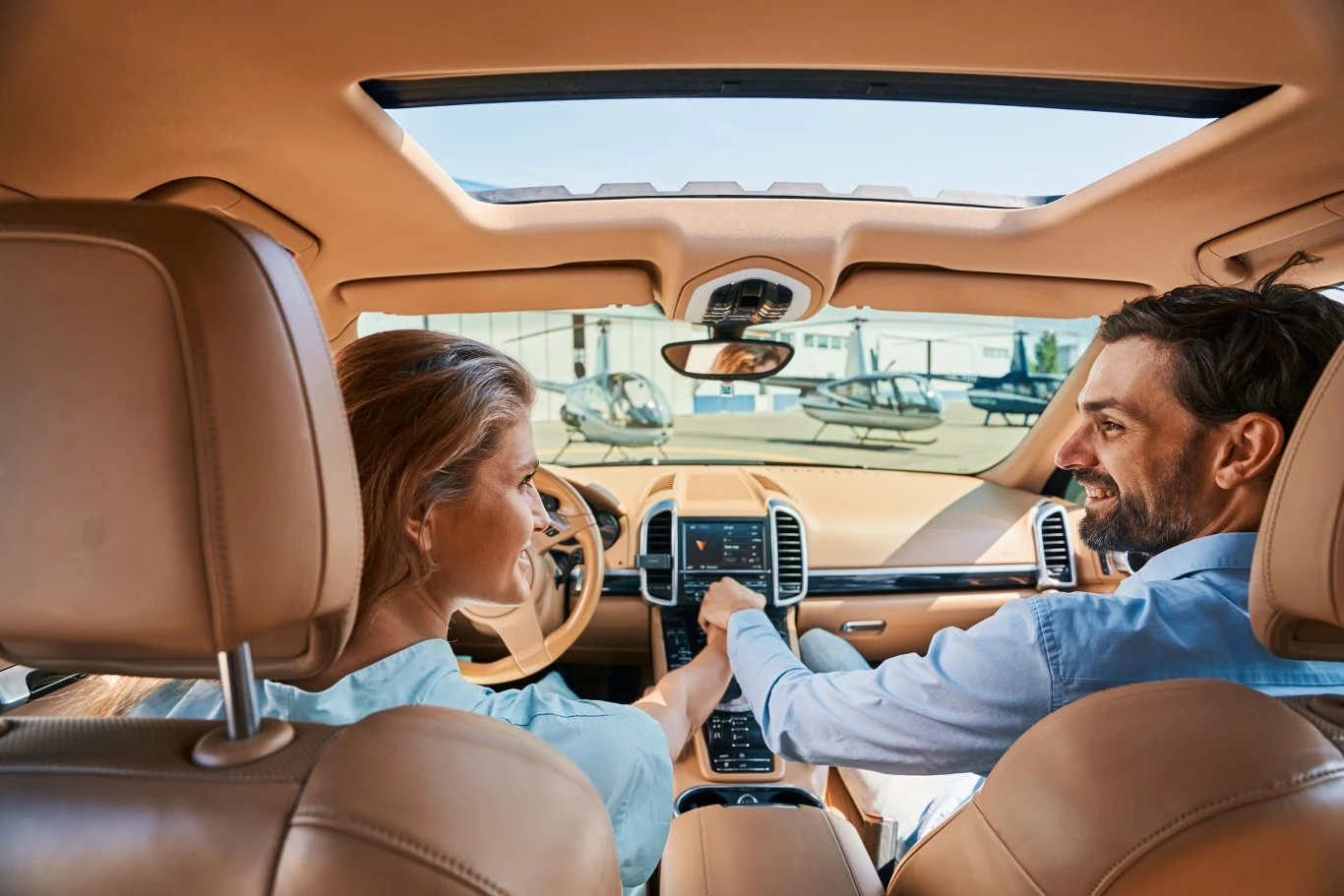 kobieta i mężczyzna siedzą w luksusowym samochodzie, uśmiechają się do siebie, wokół jasne wnętrze auta, przez okno dachowe widać niebo a przez przednią szybę helikoptery zaparkowane na tle budynków lotniska