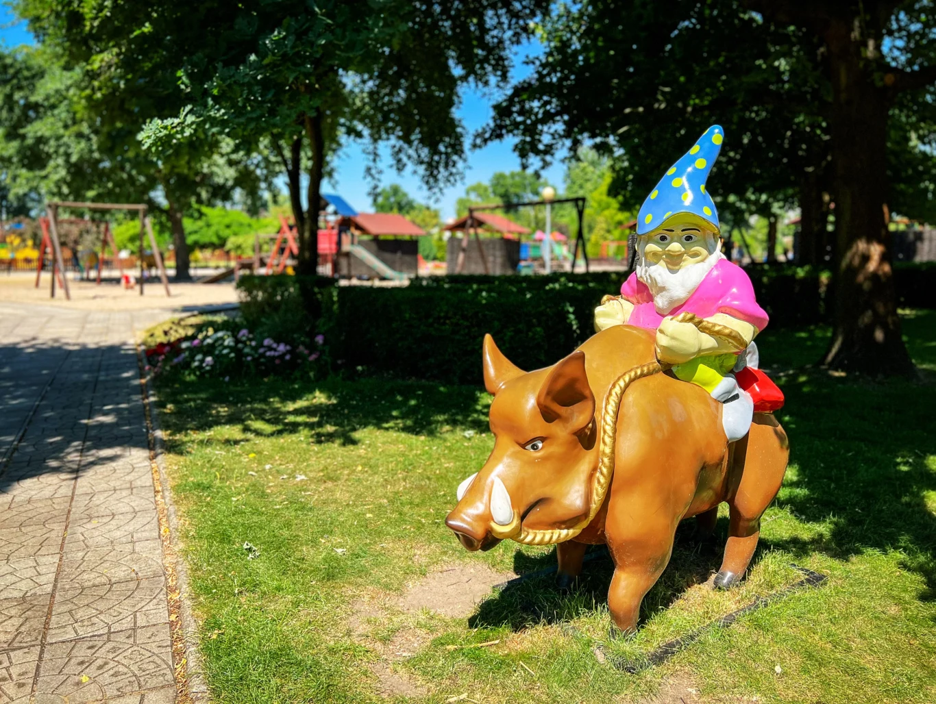 Kolorowa figura ogrodowego krasnala w niebieskiej czapce w żółte gwiazdki i różowej kurtce siedzącego na dużym dziku, ustawiona na zielonym trawniku w parku, w tle plac zabaw oraz drzewa rzucające cień.