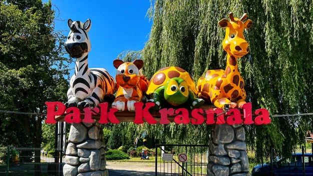 Wejście do parku ozdobione dużymi, kolorowymi figurami zwierząt: zebry, psa, żółwia i żyrafy, na kamiennych filarach, z widocznym napisem Park Krasnala i zieloną roślinnością w tle.