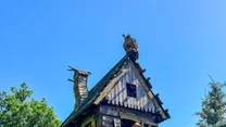 Drewniany domek stojący na rzeźbionych, stylizowanych na kurze łapy podporach, z figurką postaci bajkowej w oknie, otoczony przez ogrodzenie i drzewa iglaste, całość na tle błękitnego nieba.
