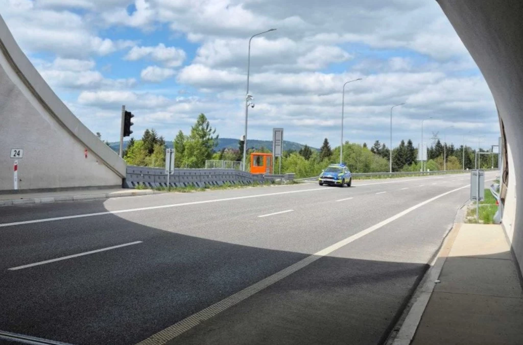 Tunel na Zakopiance to konstrukcja o długości 2058 metrów. Jasny dzień, ruchliwa droga z jednym samochodem osobowym jadącym pod wiaduktem, w tle zieleń, las i lekko pochmurne niebo, po lewej stronie widoczny sygnalizator świetlny oraz pomarańczowa skrzynka przy barierce.