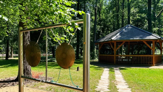 Dwa duże gongi zawieszone na metalowej ramie na tle leśnego parku, w głębi altana drewniana otoczona zielenią i ścieżką z kamiennych płyt prowadzącą do altany.
