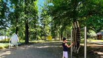 Chłopiec w ciemnej koszulce i jasnych spodenkach stoi przy nowoczesnym urządzeniu edukacyjnym w otoczeniu zielonych drzew w parku, w tle widać inne interaktywne instalacje oraz alejkę.