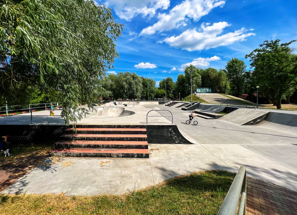 Skatepark Nowa Sól Rozległy skatepark z betonowymi rampami, poręczami i schodami, w centrum młody chłopiec jeździ na rowerze BMX, wokół widoczne drzewa oraz niebo z nielicznymi chmurami.