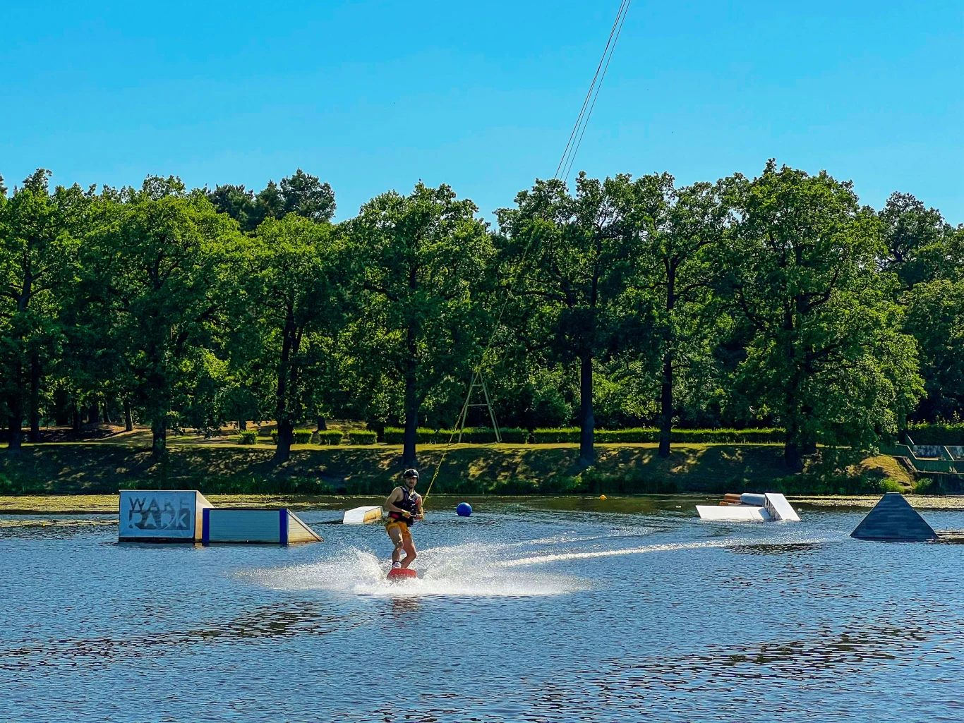 Osoba uprawiająca wakeboarding na jeziorze, otoczona przeszkodami do sportów wodnych, w tle gęsty, zielony las pod błękitnym niebem.