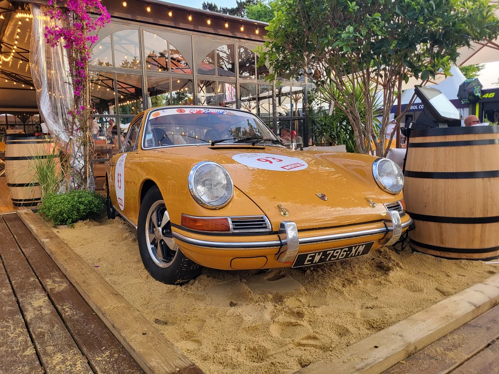 Na torze i w okolicach można spotkać mnóstwo klasycznych samochodów. Na zdjęciu - dekoracja jednego z licznych barów. Klasyczny żółty samochód sportowy Porsche stoi zaparkowany na piasku przy stylowo urządzonym tarasie restauracyjnym, otoczonym kwiatami, roślinami oraz drewnianymi beczkami, w tle widoczne szklane ściany budynku.
