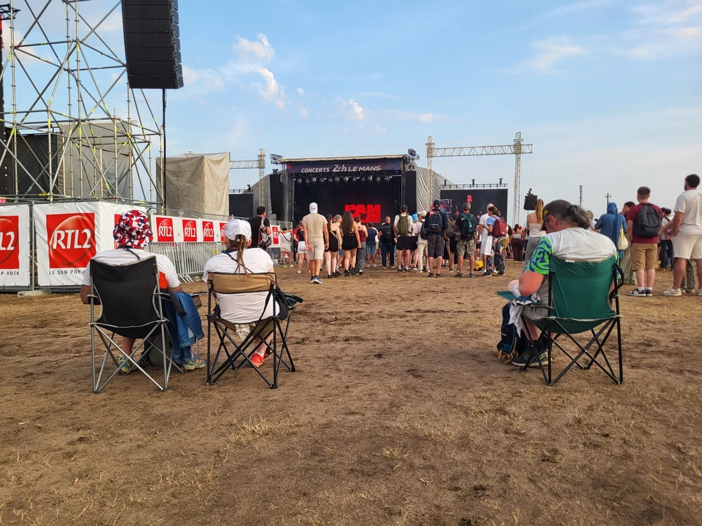 Wygodne, rozkładane krzesełka - wyposażona w nie jest większość kibiców. Scena plenerowa festiwalu muzycznego, na pierwszym planie kilka osób siedzących na składanych krzesłach, w tle scena koncertowa z widocznym tłumem oraz konstrukcjami technicznymi i banerami reklamowymi.