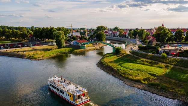 Prom płynący po rzece, otoczony zielonymi brzegami, zabudowania miejskie w tle, kilka osób na pokładzie podziwiających widoki, jasne niebo z lekkim zachmurzeniem.