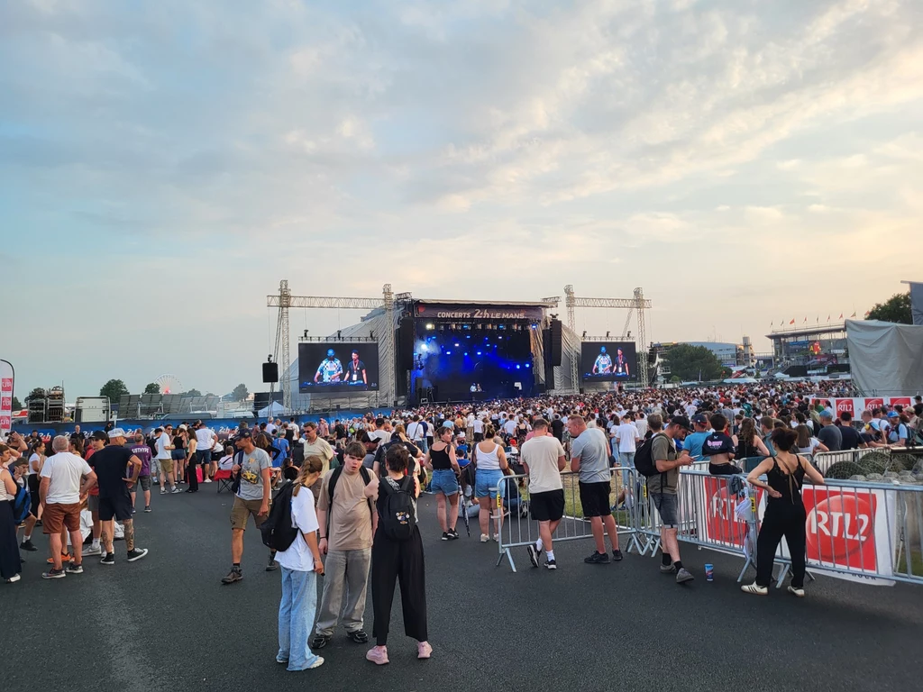 Na terenie Le Mans nie brakuje atrakcji - tu część festiwalowa, gdzie od wczesnego wieczoru odbywają się koncerty. Tłum ludzi zgromadzony pod dużą sceną koncertową na otwartym powietrzu, scena dobrze oświetlona, w tle ekrany wyświetlające artystów, wokół zabezpieczenia oraz bannery z nazwą wydarzenia, niebo z lekkim zachmurzeniem