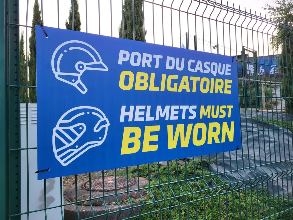 Le Mans to mekka wyścigów, o odpowiednim ubiorze przypominają tablice. Tablica informacyjna w dwóch językach, francuskim i angielskim, zawieszona na metalowym ogrodzeniu, z napisem nakazującym obowiązkowe noszenie kasków, z rysunkami kasków ochronnych.