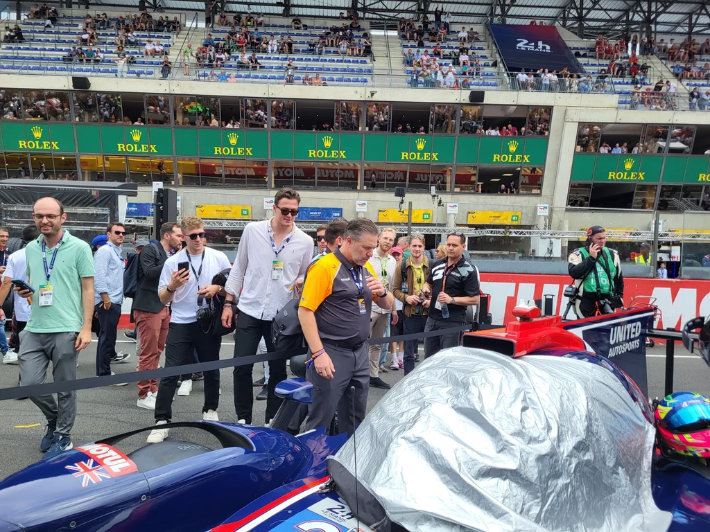 Podczas grid walku na torze można spotkać wiele znanych osób ze świata sportów motorowych - tu Zak Brown uważnie doglądający auta, które za chwilę wystartuje w wyścigu. Ludzie zgromadzeni wokół samochodu wyścigowego na torze, niektórzy fotografują i rozmawiają, w tle widownia zapełniona kibicami oraz zielone bannery z logo Rolex.