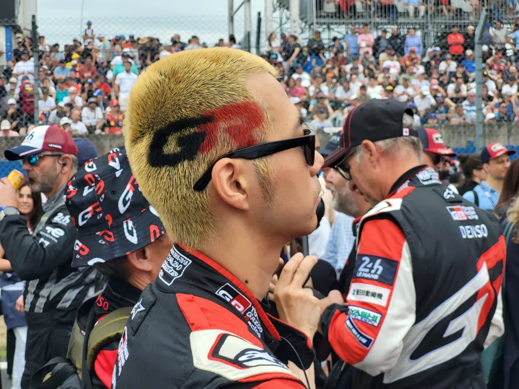 Wyścig w Le Mans to coś więcej niż zawody; frajdę będa mieli tu nawet ci, którzy niespecjalnie śledzą sporty motorowe. Mężczyzna z krótkimi blond włosami ozdobionymi wzorem w kształcie litery G i czerwonym akcentem, w okularach przeciwsłonecznych, stoi w otoczeniu zespołu wyścigowego ubranych w sportowe kombinezony, w tle liczna publiczność za ogrodzeniem.