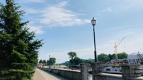Deptak wyłożony kostką prowadzący wzdłuż nabrzeża, po lewej stronie wysokie zielone drzewa, po prawej stronie murowane ogrodzenie z lampami oraz widoczna rzeka z zacumowanymi łodziami, w oddali żółty żuraw i zabudowania.