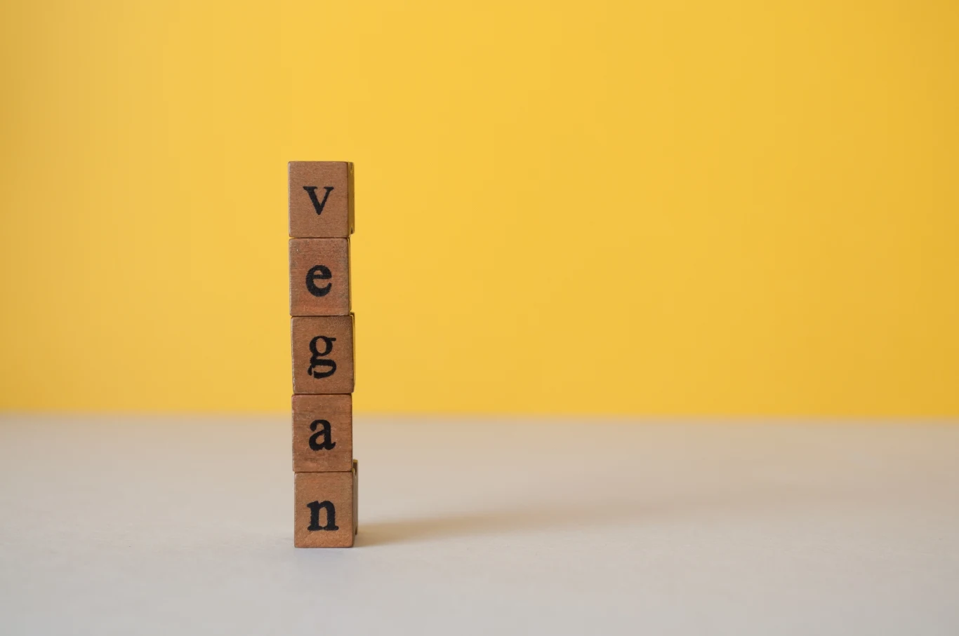 Klocki ułożone jeden na drugim tworzące pionowy napis 'vegan' na tle żółtej ściany i jasnej powierzchni