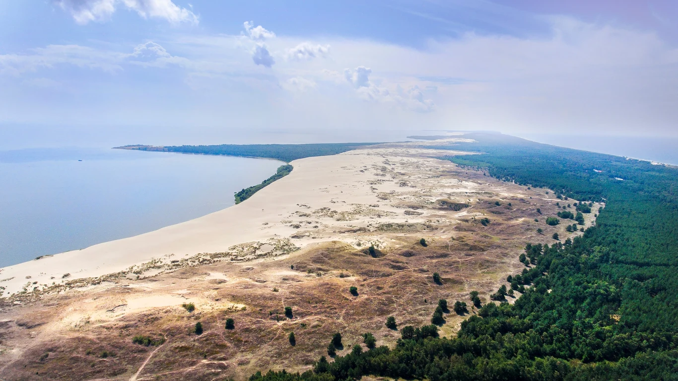 Mierzeja Kurońska, fot. Domantas Kancleris, Lithuania Travel Panorama szerokiej, piaszczystej mierzei otoczonej z jednej strony lasem, a z drugiej wodą, z wyraźnym kontrastem między jasnym piaskiem a zielenią drzew.