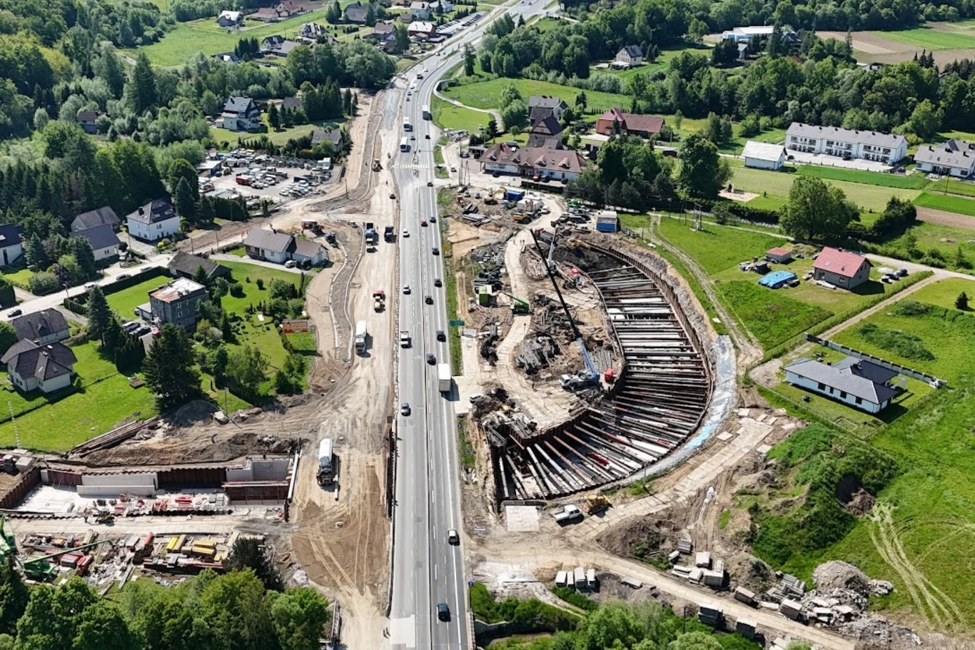 Rozległy plac budowy przy głównej drodze z nowo powstającym wiaduktem, otoczony samochodami i maszynami budowlanymi, w tle zielone tereny i zabudowa jednorodzinna.