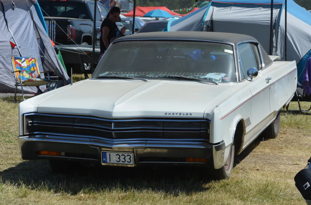 Kremowy klasyczny samochód marki Chrysler z lat 60. z czarnym dachem, zaparkowany na trawie wśród namiotów, wokół widoczni ludzie oraz biwakowy klimat.