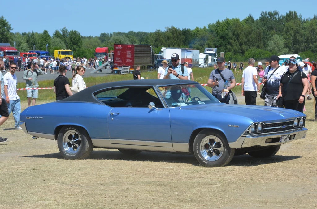Kultowy, niebieski klasyczny samochód Chevrolet Chevelle SS zaparkowany na otwartym terenie podczas wydarzenia plenerowego, wokół którego zgromadzeni są ludzie zwracający uwagę na pojazd, w tle liczne ciężarówki oraz drzewa.