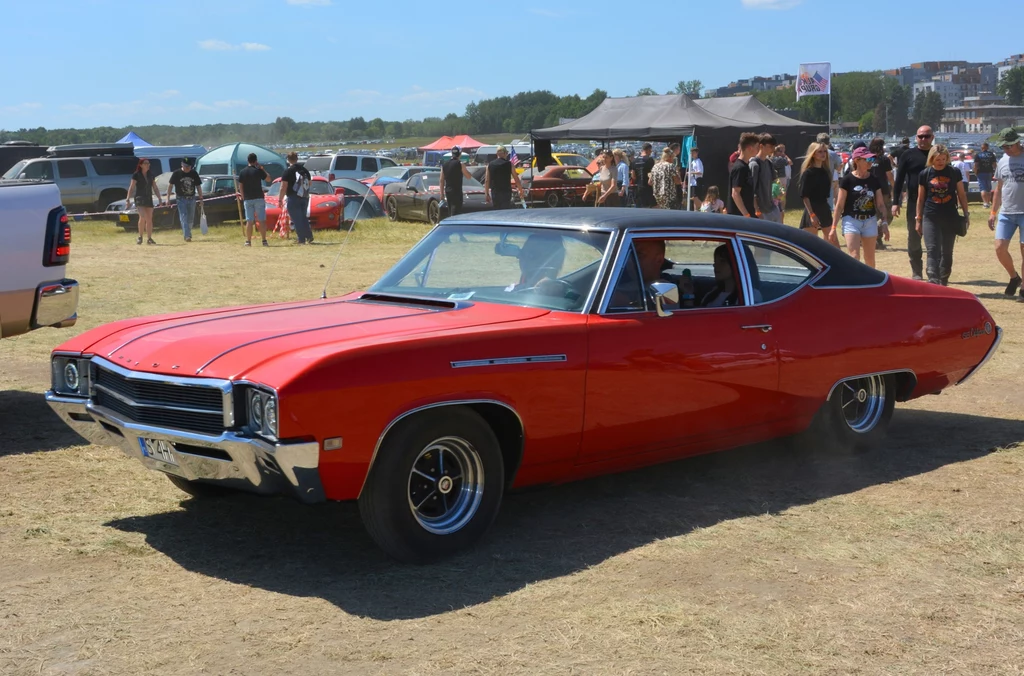 Czerwony klasyczny samochód typu muscle car na tle letniej imprezy plenerowej, otoczony przez grupę ludzi oraz zaparkowane pojazdy.