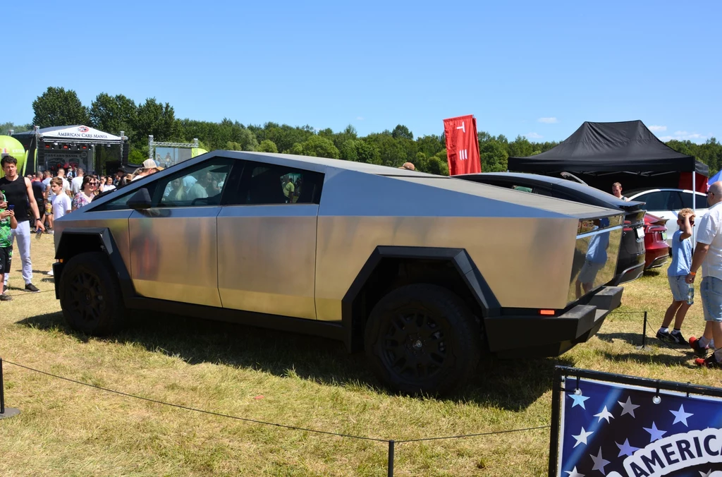 Futurystyczny samochód Tesla Cybertruck zaparkowany na otwartej przestrzeni, pośród grupy ludzi podczas wydarzenia motoryzacyjnego, na tle zielonych drzew i namiotów wystawienniczych.