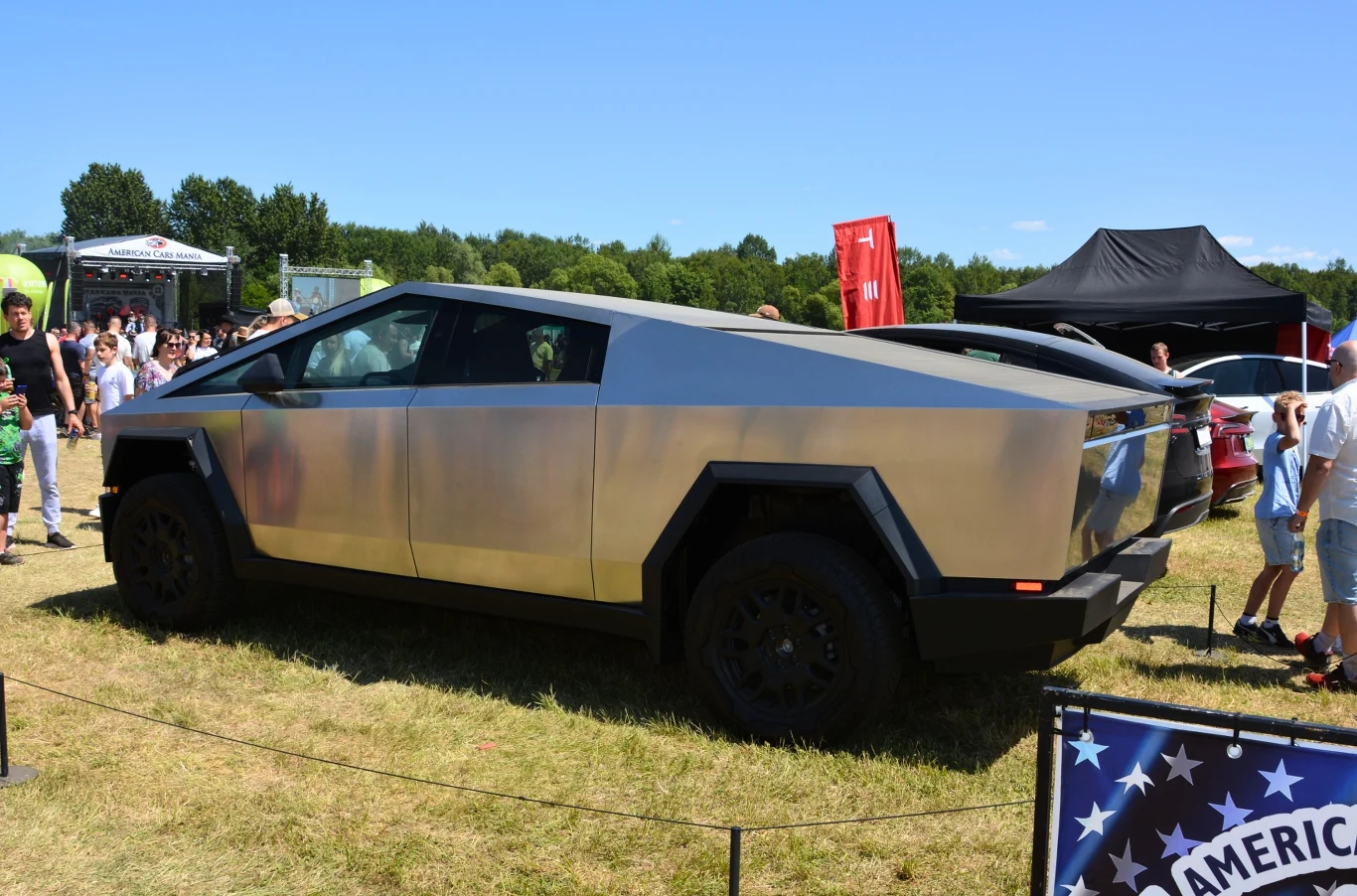 Futurystyczny samochód Tesla Cybertruck zaparkowany na otwartej przestrzeni, pośród grupy ludzi podczas wydarzenia motoryzacyjnego, na tle zielonych drzew i namiotów wystawienniczych.