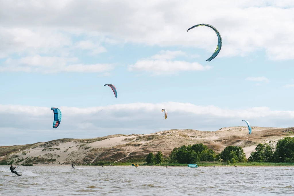 Kitesurfing w Nidzie, w tle ruchome wydmy, fot. Andrius Aleksandravičius, Lithuania Travel Kolorowe latawce do kitesurfingu unoszące się nad wodą, poniżej kilku sportowców uprawiających kitesurfing, w tle piaszczyste wydmy i zieleń drzew pod niebieskim, lekko zachmurzonym niebem.
