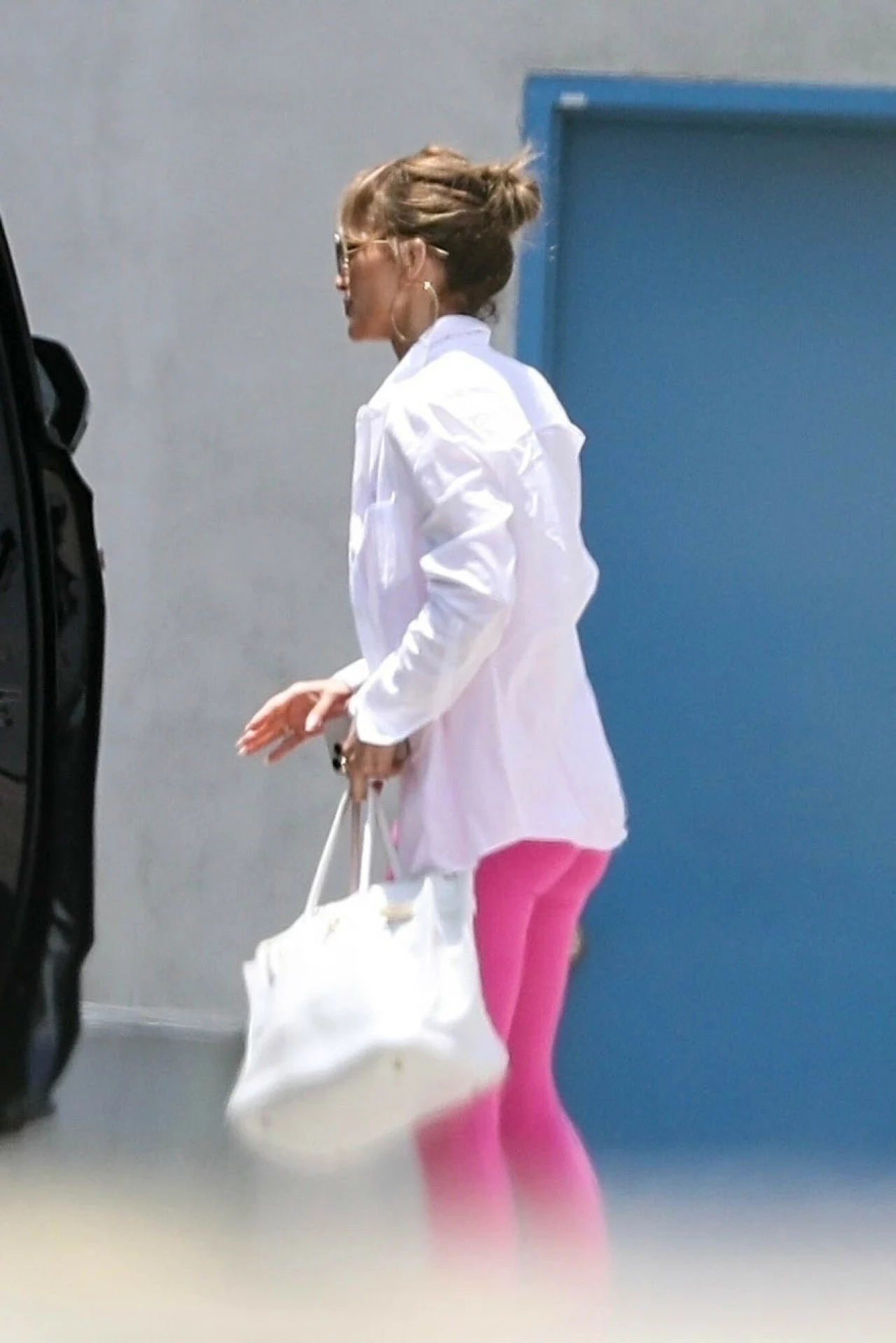 Jennifer Lopez przygotowuje się do trasy koncertowej Kobieta w białej koszuli i różowych legginsach trzymająca białą torbę, idąca obok jasnej ściany i niebieskich drzwi.
