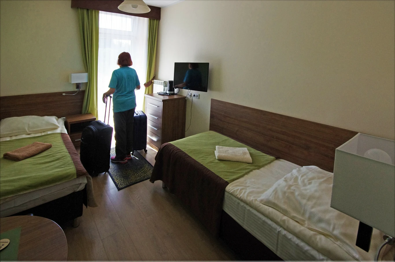 Dobrze spakowana sanatoryjna walizka to gwarancja udanego pobytu Kobieta z walizką stoi przy oknie w jasno oświetlonym pokoju hotelowym z dwoma pojedynczymi łóżkami, zielonymi i brązowymi narzutami oraz telewizorem zawieszonym na ścianie.