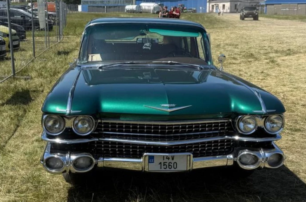Klasyczny, zielony samochód marki Cadillac z lat 50. lub 60., sfotografowany od frontu, stoi na trawiastym terenie, w tle widoczne są inne pojazdy oraz zabudowania przemysłowe.