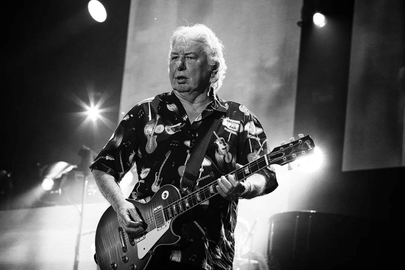 Starszy mężczyzna o siwych włosach gra na gitarze elektrycznej na scenie koncertowej, ubrany w koszulę z motywami gitar, wokół intensywne oświetlenie sceniczne.