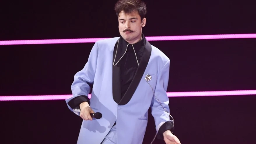 Wiktor Dyduła Mężczyzna z wąsem w jasno-niebieskim garniturze i czarnej koszuli z ozdobnym łańcuszkiem, trzymający mikrofon, stoi na scenie oświetlonej różowymi liniami świetlnymi.