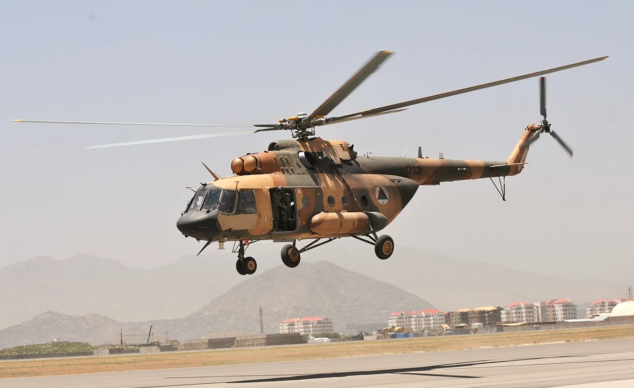 Wojskowy helikopter w barwach kamuflażu unosi się nad pasem startowym, w tle widoczne są zabudowania miejskie oraz górzysty krajobraz.