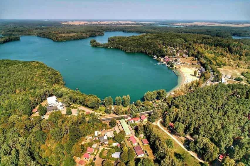 Rozległe jezioro otoczone gęstymi lasami, z zamieszkałą strefą domków letniskowych, plażą oraz infrastrukturą rekreacyjną, na horyzoncie rozległe przestrzenie leśne i wodne.