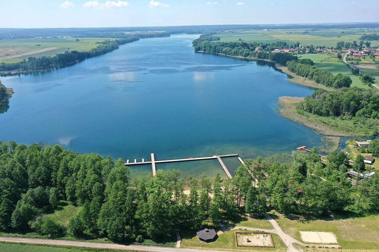 Rozległe jezioro otoczone zielonymi polami i lasami, z widocznym pomostem nad wodą oraz piaszczystą plażą po jednej stronie brzegu, w tle widać zabudowania wiejskie.