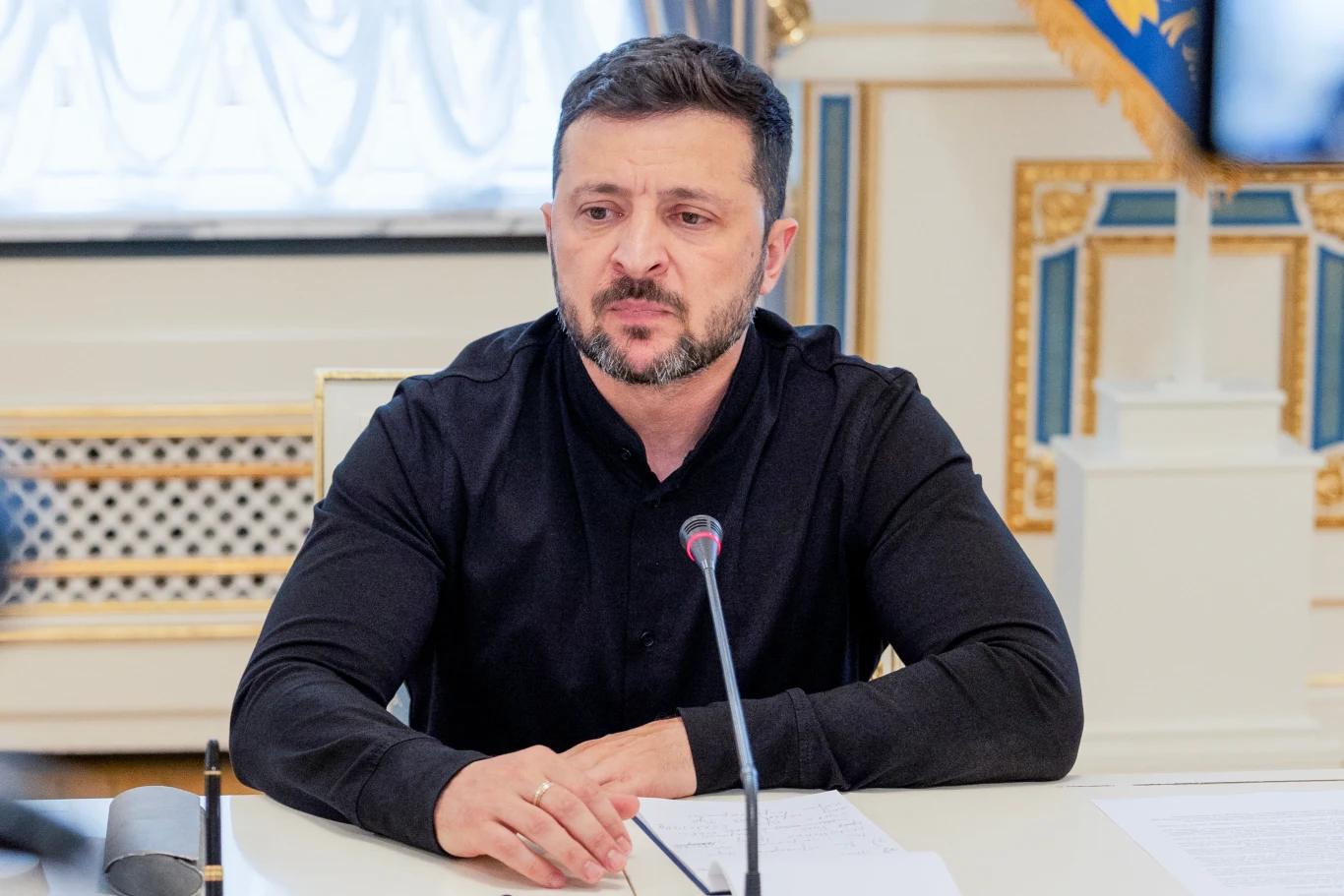 Wołodymyr Zełenski Mężczyzna w ciemnej koszuli siedzi przy stole z mikrofonem, przed nim leżą dokumenty, w tle eleganckie wnętrze z zasłonami i ozdobami.